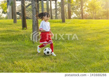 Active little girl in red and white uniform running after a soccer ball on green grass hill. Active little girl in red and white uniform running after a soccer ball on green grass hill. 141688266