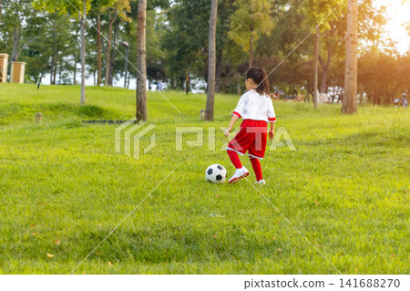 Active little girl in red and white uniform running after a soccer ball on green grass hill. Active little girl in red and white uniform running after a soccer ball on green grass hill. 141688270