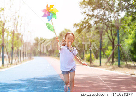 Cheerful little girl running forward while holding a colorful pinwheel toy on a park path. 141688277