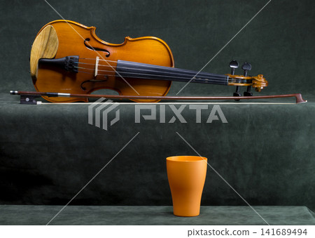 Still life with a violin, bow, and orange cup on a green background. Still life with a violin, bow, and orange cup on a green background. 141689494