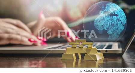 Stacked gold bullion bars and digital wireframe Earth globe on desk with blurred woman typing on laptop in background. 141689501
