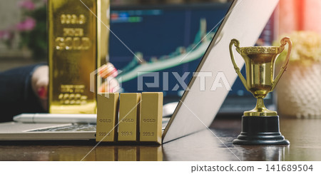 Financial achievement. Gold bars and trophy next to a laptop with blurred trading graphs in the background. Financial achievement. Gold bars and trophy next to a laptop with blurred trading graphs in the background. 141689504
