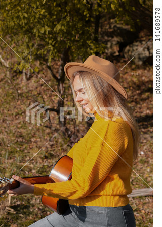 Beautiful blonde girl playing acoustic guitar and singing relaxing enjoying outdoors on sunny autumn day. Hippie woman in boho style learning and practicing playing musical instrument. Fall leaves Beautiful blonde girl playing acoustic guitar and singing relaxing enjoying outdoors on sunny autumn day. Hippie woman in boho style learning and practicing playing musical instrument. Fall leaves 141689578
