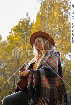 Beautiful blonde girl playing acoustic guitar and singing relaxing enjoying outdoors on sunny autumn day. Hippie woman in boho style learning and practicing playing musical instrument. Fall leaves 141689584