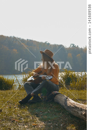 Beautiful blonde girl playing acoustic guitar and singing relaxing enjoying outdoors on sunny autumn day. Hippie woman in boho style learning and practicing playing musical instrument. Fall leaves Beautiful blonde girl playing acoustic guitar and singing relaxing enjoying outdoors on sunny autumn day. Hippie woman in boho style learning and practicing playing musical instrument. Fall leaves 141689586