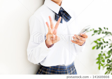 A female middle school student in a school uniform holds up three fingers while holding up a smartphone. A female middle school student in a school uniform holds up three fingers while holding up a smartphone. 141689808