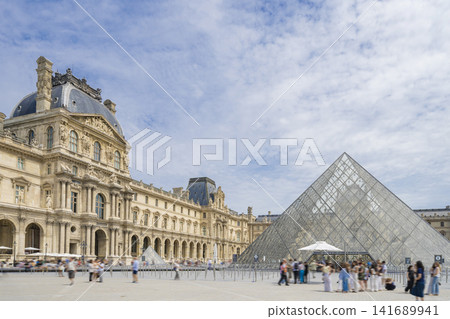 The Louvre Museum and the glass pyramid against a backdrop of blue skies and clouds. 141689941