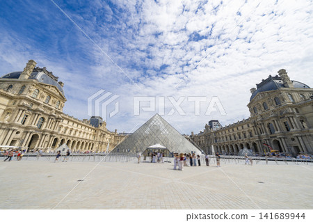 The Louvre Museum and the glass pyramid against a backdrop of blue skies and clouds. 141689944