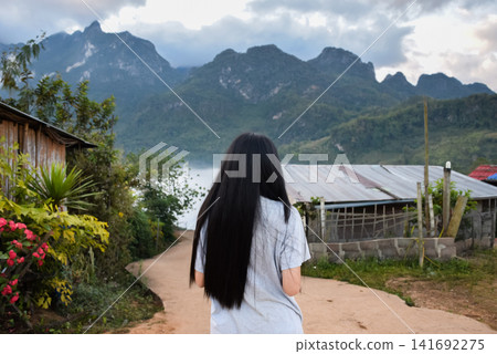 Woman standing rural road facing misty mountains village homes under dramatic clouds peaceful travel destination scenic nature outdoor lifestyle atmosphere Woman standing rural road facing misty mountains village homes under dramatic clouds peaceful travel destination scenic nature outdoor lifestyle atmosphere 141692275