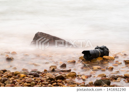 Professional camera resting pebbles beside calm water captured long exposure creating smooth motion ideal for travel photography nature landscape outdoor themes Professional camera resting pebbles beside calm water captured long exposure creating smooth motion ideal for travel photography nature landscape outdoor themes 141692290