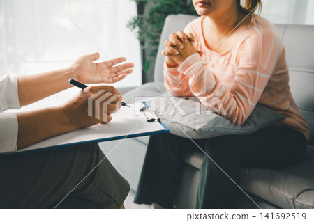 Female client listens with clasped hands as therapist gestures during counseling, showing private consultation, mental health support, empathy, guidance, stress relief, and trust indoors. 141692519