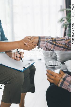 Female client shakes therapist hand while holding water after counseling on sofa, showing gratitude, trust, mental health support, reassurance, professional care, and emotional healing indoors. Female client shakes therapist hand while holding water after counseling on sofa, showing gratitude, trust, mental health support, reassurance, professional care, and emotional healing indoors. 141692521