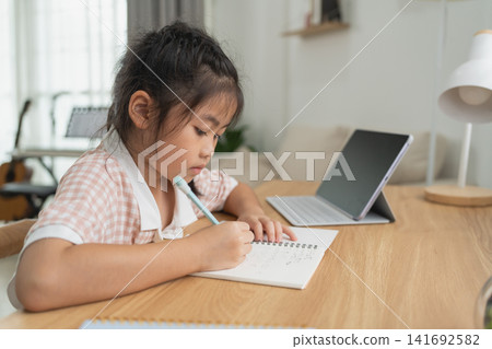 Young girl focused on writing in notebook with pen while studying at wooden table with laptop and lamp Young girl focused on writing in notebook with pen while studying at wooden table with laptop and lamp 141692582