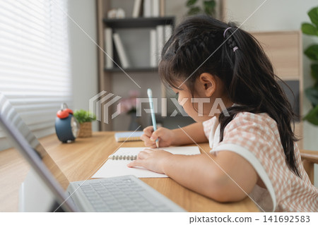 Young girl focused on writing in notebook while sitting at wooden desk with laptop in cozy home setting Young girl focused on writing in notebook while sitting at wooden desk with laptop in cozy home setting 141692583