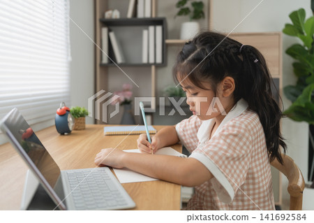 Young girl focused on writing homework sitting at wooden desk with laptop and plant in bright home study room Young girl focused on writing homework sitting at wooden desk with laptop and plant in bright home study room 141692584