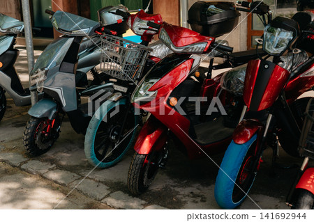 Red and gray electric scooters stand parked in a row on a city sidewalk. Personal transport, electric mobility, urban commuting, and modern transport solutions Red and gray electric scooters stand parked in a row on a city sidewalk. Personal transport, electric mobility, urban commuting, and modern transport solutions 141692944