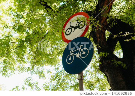 A bicycle lane sign stands below a moped restriction sign among green tree branches. Urban traffic control, sustainable transport, safety, and eco mobility 141692956