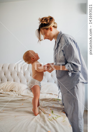 Mother helping baby stand up on bed during morning routine Mother helping baby stand up on bed during morning routine 141693021