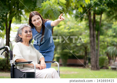Caucasian female caregiver encouraging senior patient on wheelchair in garden. Caucasian female caregiver encouraging senior patient on wheelchair in garden. 141693415