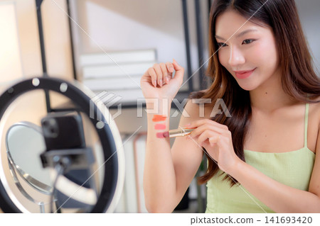Young asian beauty influencer testing multiple lipstick shades on arm while recording makeup review. Young asian beauty influencer testing multiple lipstick shades on arm while recording makeup review. 141693420