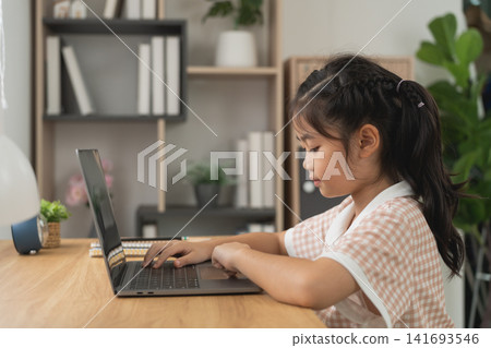 Young Asian girl focused on using laptop computer at wooden table in modern cozy home environment 141693546