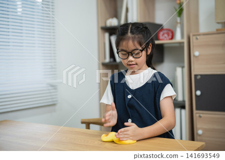 Asian Little Girl with Glasses Squeezing Yellow Squishy Toys with Both Hands on Wooden Desk in Home Workspace for Fine Motor Skills Practice Asian Little Girl with Glasses Squeezing Yellow Squishy Toys with Both Hands on Wooden Desk in Home Workspace for Fine Motor Skills Practice 141693549