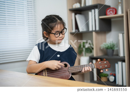 Cute Asian Little Girl with Glasses Focused on Playing Ukulele at Wooden Desk in Bright Modern Home Living Room Workspace Cute Asian Little Girl with Glasses Focused on Playing Ukulele at Wooden Desk in Bright Modern Home Living Room Workspace 141693550