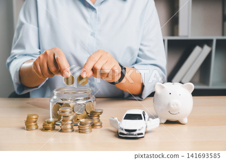 Person putting coins into a glass jar with piggy bank and white toy car on desk, representing saving money for car purchase, down payment, auto loan planning, and vehicle finance. Person putting coins into a glass jar with piggy bank and white toy car on desk, representing saving money for car purchase, down payment, auto loan planning, and vehicle finance. 141693585