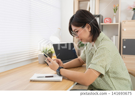 Asian woman using smartphone while sitting at wooden desk with laptop in bright minimalist home office workspace Asian woman using smartphone while sitting at wooden desk with laptop in bright minimalist home office workspace 141693675