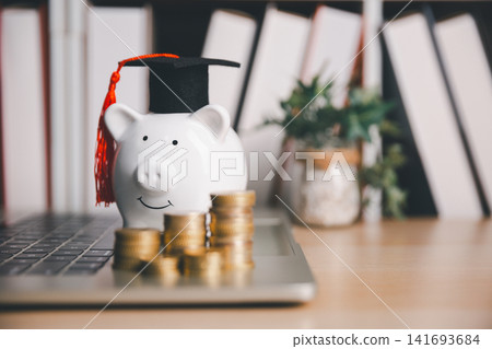 Piggy bank wearing a graduation cap on a laptop with stacked coins, representing college savings, online tuition payment, digital financial planning, and investment in education. 141693684