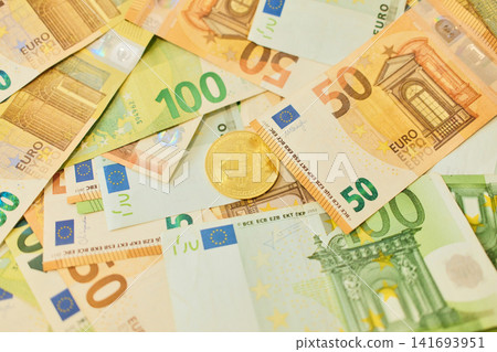 A gold bitcoin token lies on mixed 50 and 100 euro banknotes in a layered money background. Digital currency and finance concept combining crypto and traditional cash A gold bitcoin token lies on mixed 50 and 100 euro banknotes in a layered money background. Digital currency and finance concept combining crypto and traditional cash 141693951