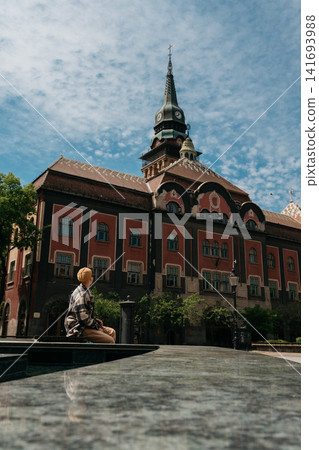 Traveler young woman sits by the fountain facing Subotica City Hall in Subotica, Serbia. Solo travel, urban exploration, and peaceful tourism in a historic European city Traveler young woman sits by the fountain facing Subotica City Hall in Subotica, Serbia. Solo travel, urban exploration, and peaceful tourism in a historic European city 141693988