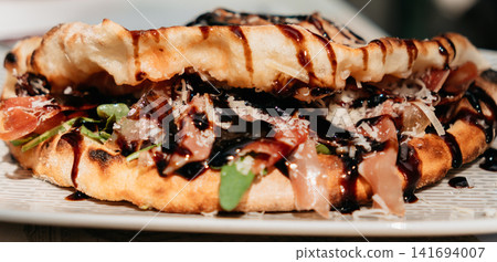 Crispy folded flatbread filled with prosciutto, greens and cheese, finished with balsamic glaze. Gourmet street food presentation creates a premium lunch concept Crispy folded flatbread filled with prosciutto, greens and cheese, finished with balsamic glaze. Gourmet street food presentation creates a premium lunch concept 141694007