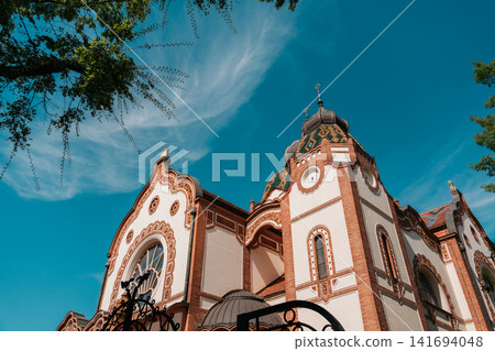 Decorative facade of Subotica Synagogue under vivid blue sky in Subotica, Serbia. Art Nouveau architecture with ornate brickwork and ceramic roof details Decorative facade of Subotica Synagogue under vivid blue sky in Subotica, Serbia. Art Nouveau architecture with ornate brickwork and ceramic roof details 141694048