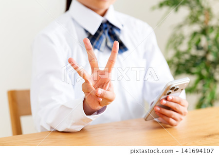 一名身穿校服的中學生坐在客廳的桌子上,看著自己的智慧型手機,並舉起了三根手指。 一名身穿校服的中學生坐在客廳的桌子上,看著自己的智慧型手機,並舉起了三根手指。 141694107