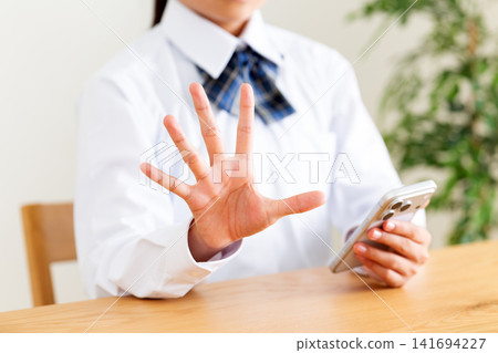 一名身穿校服的中學生坐在客廳的桌子上,看著自己的智慧型手機,並伸出了五個手指。 一名身穿校服的中學生坐在客廳的桌子上,看著自己的智慧型手機,並伸出了五個手指。 141694227