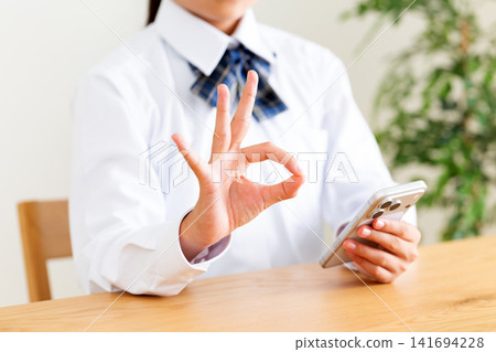 一名身穿校服的中學生正在客廳的桌子上看著她的智慧型手機,並用手指畫著圈圈。 一名身穿校服的中學生正在客廳的桌子上看著她的智慧型手機,並用手指畫著圈圈。 141694228