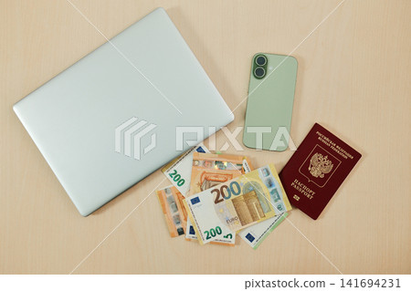 Laptop, green smartphone, euro banknotes and Russian passport are arranged on a clean desk. Concept of relocation from Russia, remote work and financial planning for moving abroad Laptop, green smartphone, euro banknotes and Russian passport are arranged on a clean desk. Concept of relocation from Russia, remote work and financial planning for moving abroad 141694231