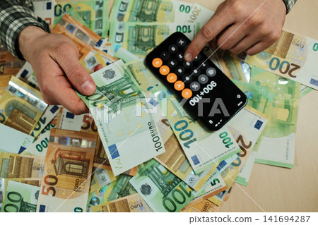 Hands hold 100 euro banknotes while calculating totals on a smartphone calculator over scattered cash. Concept of personal finance, expense control and budgeting for banking or business use Hands hold 100 euro banknotes while calculating totals on a smartphone calculator over scattered cash. Concept of personal finance, expense control and budgeting for banking or business use 141694287