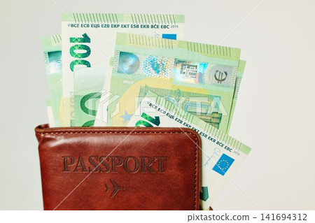 Close up of brown passport wallet with 100 euro banknotes on white background. Travel savings concept with passport holder, European cash and vacation finance Close up of brown passport wallet with 100 euro banknotes on white background. Travel savings concept with passport holder, European cash and vacation finance 141694312
