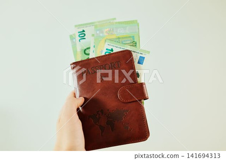 Hand holds brown passport wallet with 100 euro banknotes on white background. Travel savings concept with passport, European cash and financial planning 141694313