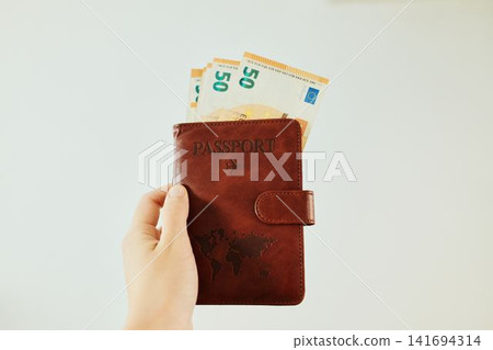 Hand holds brown passport wallet with 50 euro banknotes on white background. Travel payment concept with passport, cash and vacation budget planning Hand holds brown passport wallet with 50 euro banknotes on white background. Travel payment concept with passport, cash and vacation budget planning 141694314
