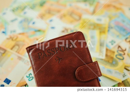 Close up of brown passport wallet on blurred euro banknotes background. Travel finance concept with passport holder, European currency and vacation budget Close up of brown passport wallet on blurred euro banknotes background. Travel finance concept with passport holder, European currency and vacation budget 141694315