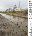 A picturesque view of a flock of ducks swimming in a shallow urban canal with an ornate church featuring golden domes in the background, surrounded by new residential high rise buildings in a city. 141694427