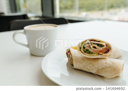 Salmon tortilla wrap with cappuccino on a bright cafe table near the window. Concept of urban breakfast, business lunch, coffee culture and restaurant branding Salmon tortilla wrap with cappuccino on a bright cafe table near the window. Concept of urban breakfast, business lunch, coffee culture and restaurant branding 141694692