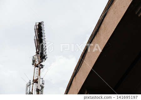 Large stadium floodlight rises beside urban structure under cloudy sky. Concept of sport infrastructure, technology and public events 141695197