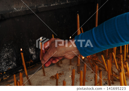 Person places a lit candle into white sand inside a religious shrine. Concept of prayer, faith, spirituality and hope Person places a lit candle into white sand inside a religious shrine. Concept of prayer, faith, spirituality and hope 141695223