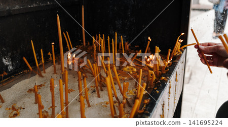 Human hand places thin candles into a religious prayer stand. Concept of faith, spirituality, remembrance and personal devotion Human hand places thin candles into a religious prayer stand. Concept of faith, spirituality, remembrance and personal devotion 141695224