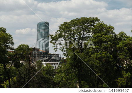 Modern glass tower rises behind green trees in Belgrade Serbia. Concept of sustainable city growth, business and urban nature Modern glass tower rises behind green trees in Belgrade Serbia. Concept of sustainable city growth, business and urban nature 141695497