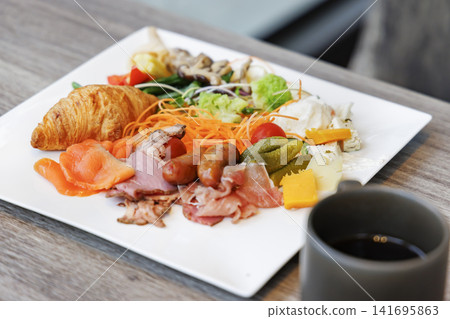 A breakfast plate generously topped with croissants, prosciutto, and other items, along with coffee. A breakfast plate generously topped with croissants, prosciutto, and other items, along with coffee. 141695863